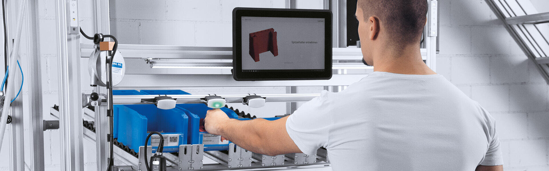 Eine Person in einem weißen Hemd scannt mit einem Handscanner blaue Behälter in einem Metallregal, während im Hintergrund ein Monitor Anweisungen anzeigt.