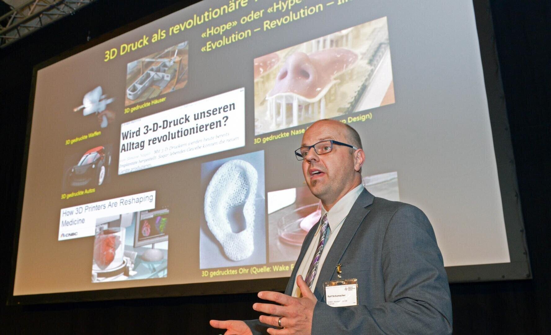 A man in a suit and glasses speaks in front of a screen showing a presentation on 3D printing in medicine, with pictures of printed organs and implants and the accompanying text in German.
