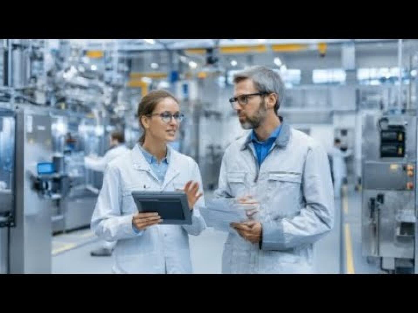 Zwei Ingenieure in Laborkitteln und Brillen besprechen ihre Arbeit, während sie ein Tablet und Dokumente in einer modernen, hell erleuchteten Industriefabrik mit Maschinen im Hintergrund halten.