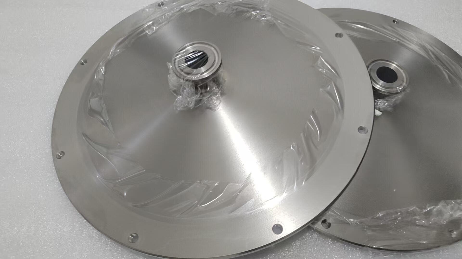 Two shiny, round metal plates with raised fittings in the center, encased in plastic and with evenly spaced holes near the edges, are placed on a white, textured surface.