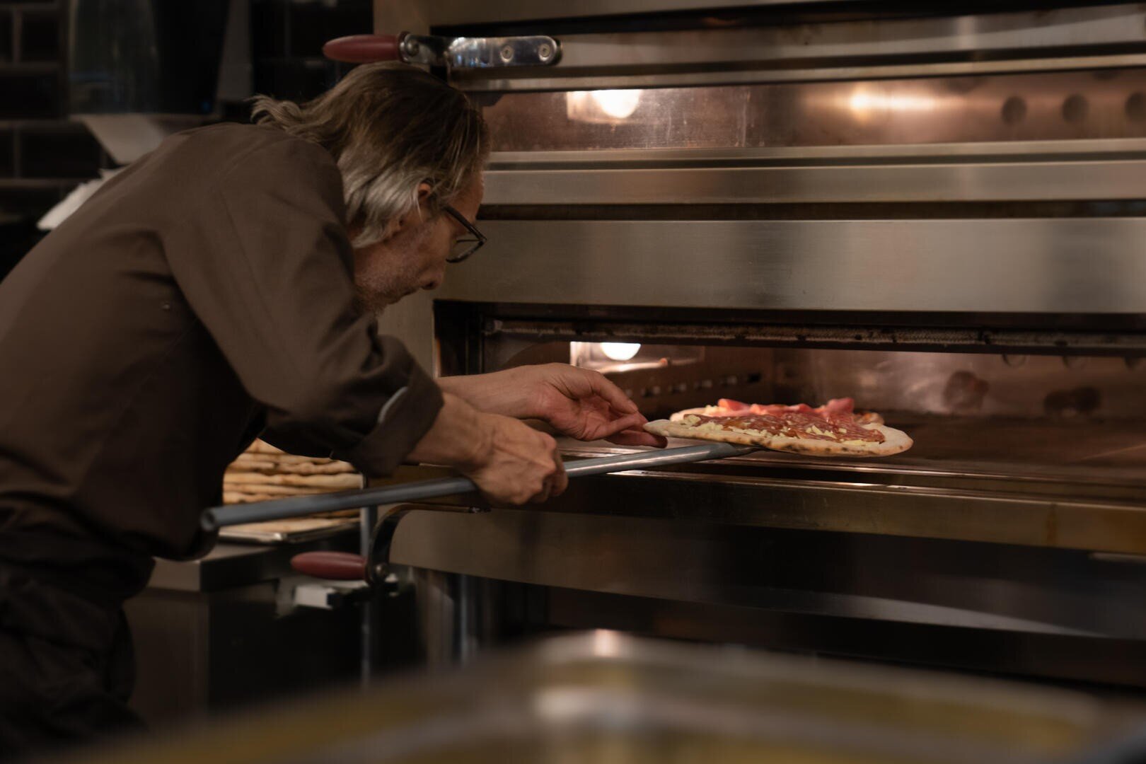 Eine Person mit grauen Haaren und Brille, die ein braunes Hemd trägt, benutzt einen Pizzaschieber, um eine Pizza mit Belag in einen großen Pizzaofen zu schieben.
