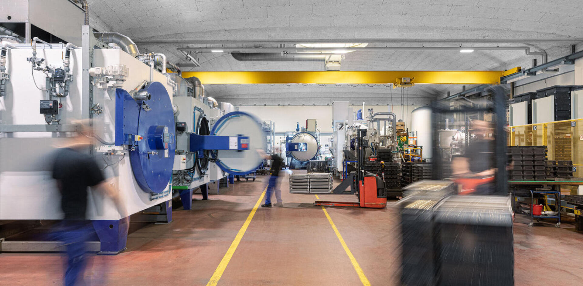 Eine moderne Industrieanlage mit großen Maschinen, unscharfen Arbeitern in Bewegung und gelben Sicherheitslinien auf dem Boden, die auf eine aktive, auf Produktion und Sicherheit ausgerichtete Fertigungsumgebung hinweisen.
