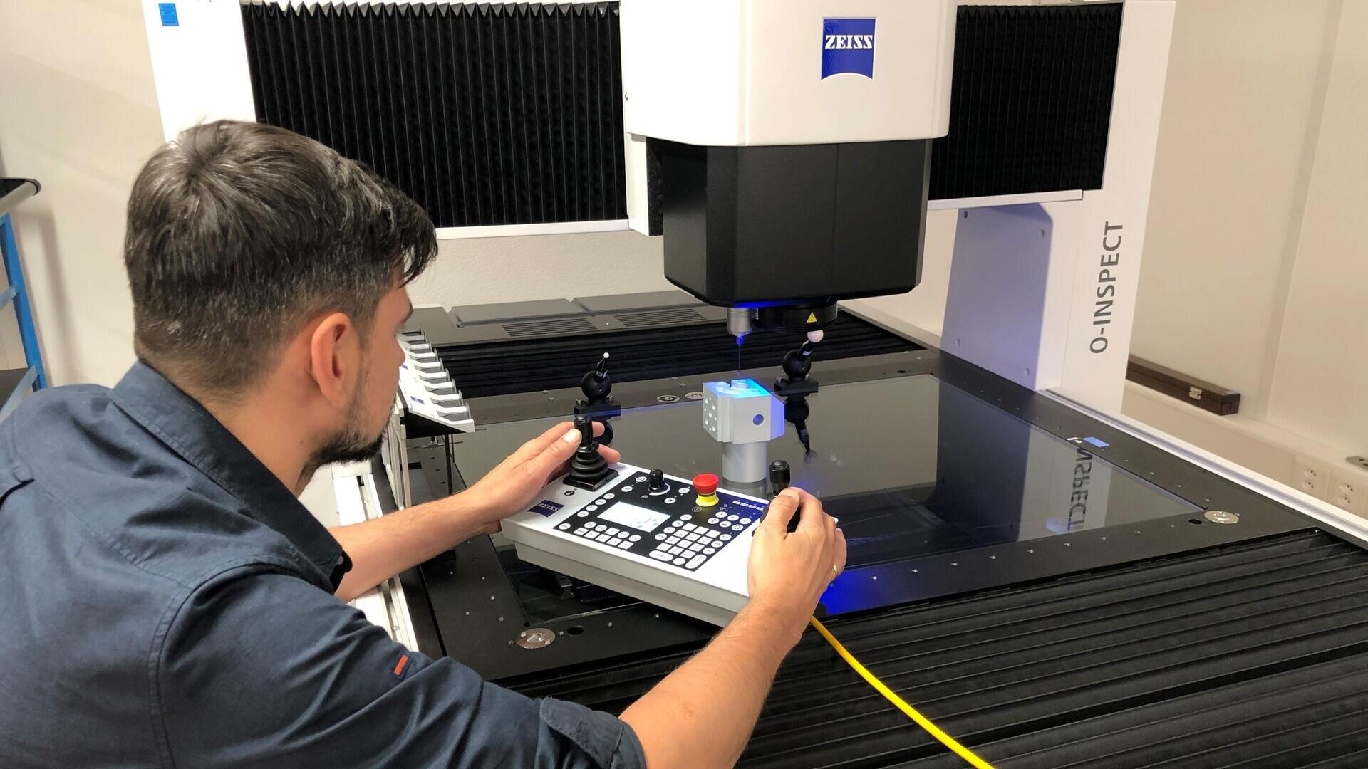 Ein Mann bedient eine ZEISS O-INSPECT-Maschine und prüft mithilfe eines Bedienfelds ein kleines Objekt, das in einer Laborumgebung auf der Glasplattform der Maschine platziert ist.