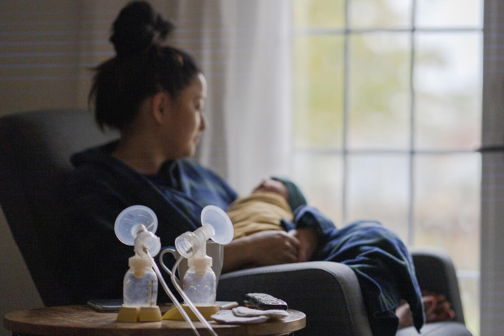 A woman sits on a chair by the window holding a swaddled baby. In the foreground is a breast pump with two feeding bottles on a small table. The scene appears calm and softly lit.