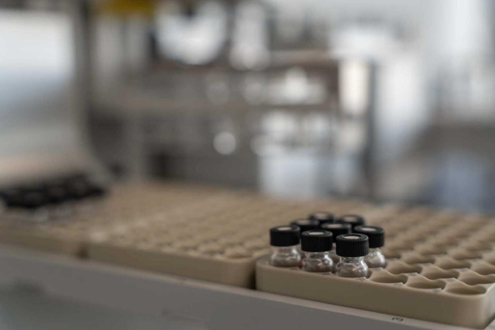 Nahaufnahme mehrerer kleiner Glasfläschchen mit schwarzen Deckeln, die in einem beigen Labortablett platziert sind, mit einem unscharfen wissenschaftlichen Laborhintergrund.