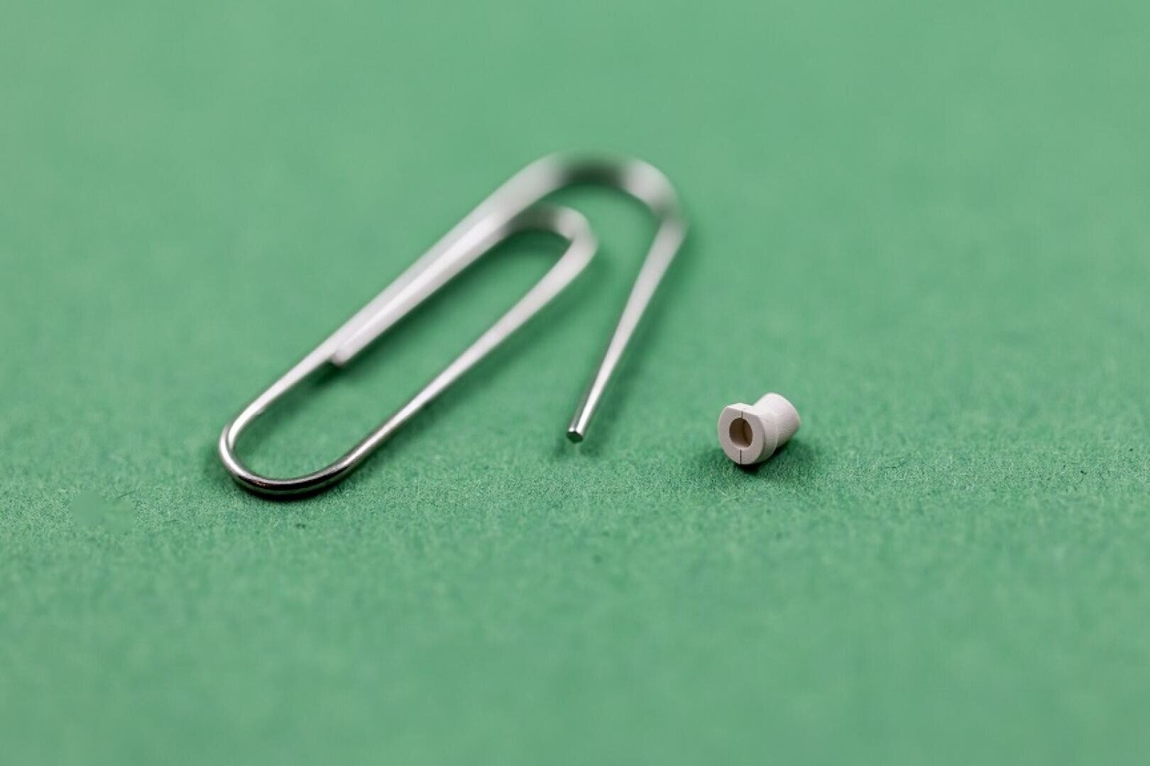 A small white, cylindrical object is placed next to a silver paper clip on a green surface to show the tiny size of the object in comparison.