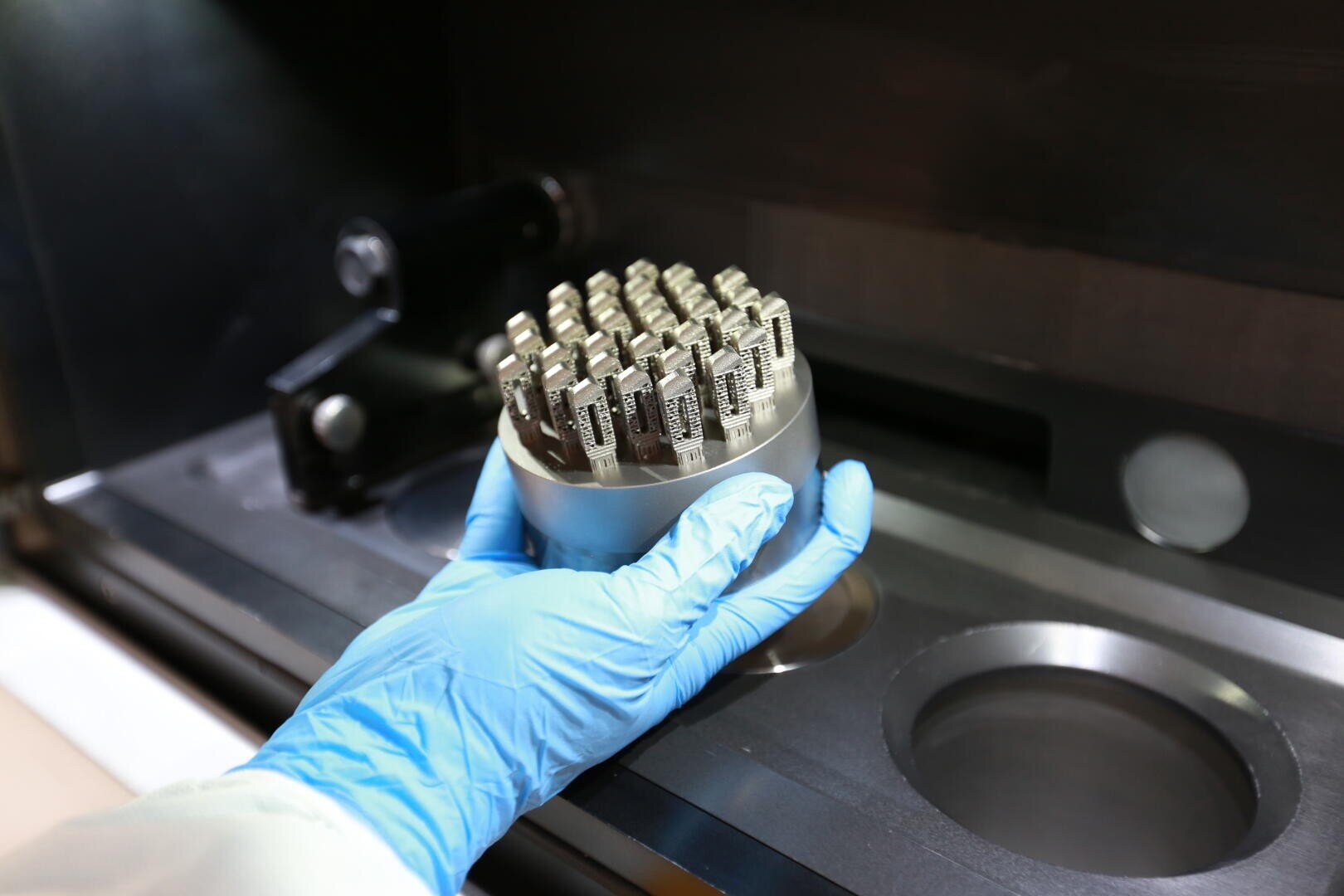 Eine behandschuhte Hand hält einen Metallbehälter, der mit zahlreichen kleinen, kompliziert geformten Metallteilen gefüllt ist, die wahrscheinlich in einem Hightech-Fertigungs- oder 3D-Druckverfahren hergestellt wurden.