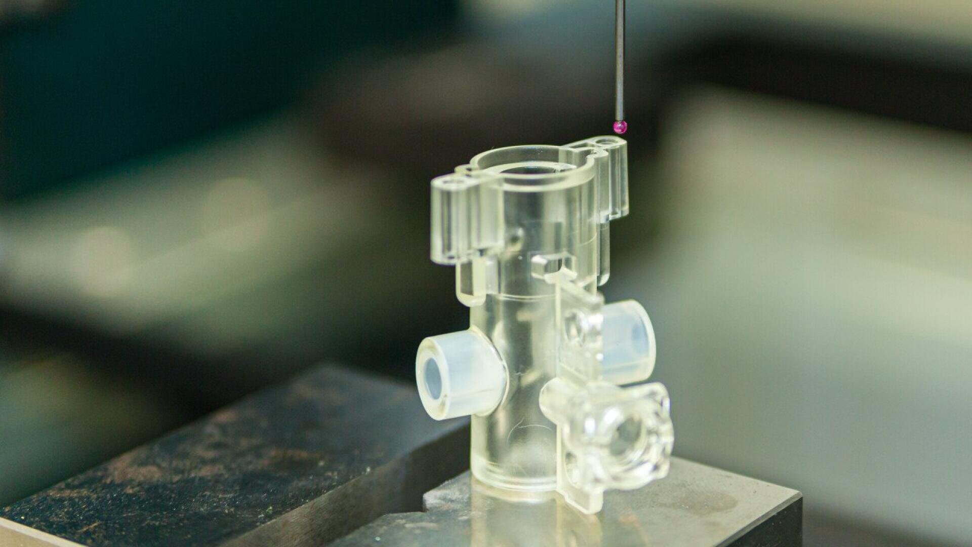 A clear, cylindrical plastic component is being measured with a precision probe on a metallic surface, indicating a quality control or inspection process in a laboratory or manufacturing setting.