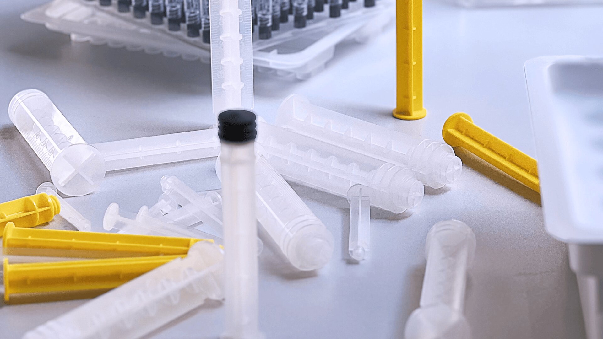 Mehrere durchsichtige Plastikspritzen und gelbe Plastikröhrchen liegen verstreut auf einer weißen Fläche, im Hintergrund sind weitere verpackte Spritzen und medizinisches Zubehör zu sehen.