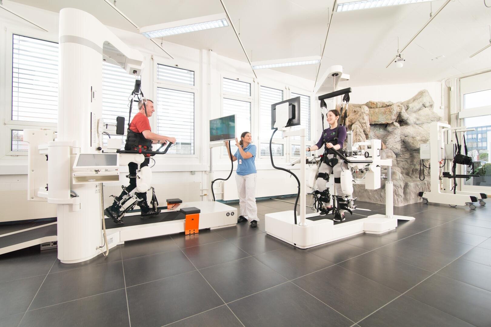In a bright, modern clinic with large windows and stone walls, two people use a robotic exoskeleton for rehabilitation therapy while a therapist monitors them.