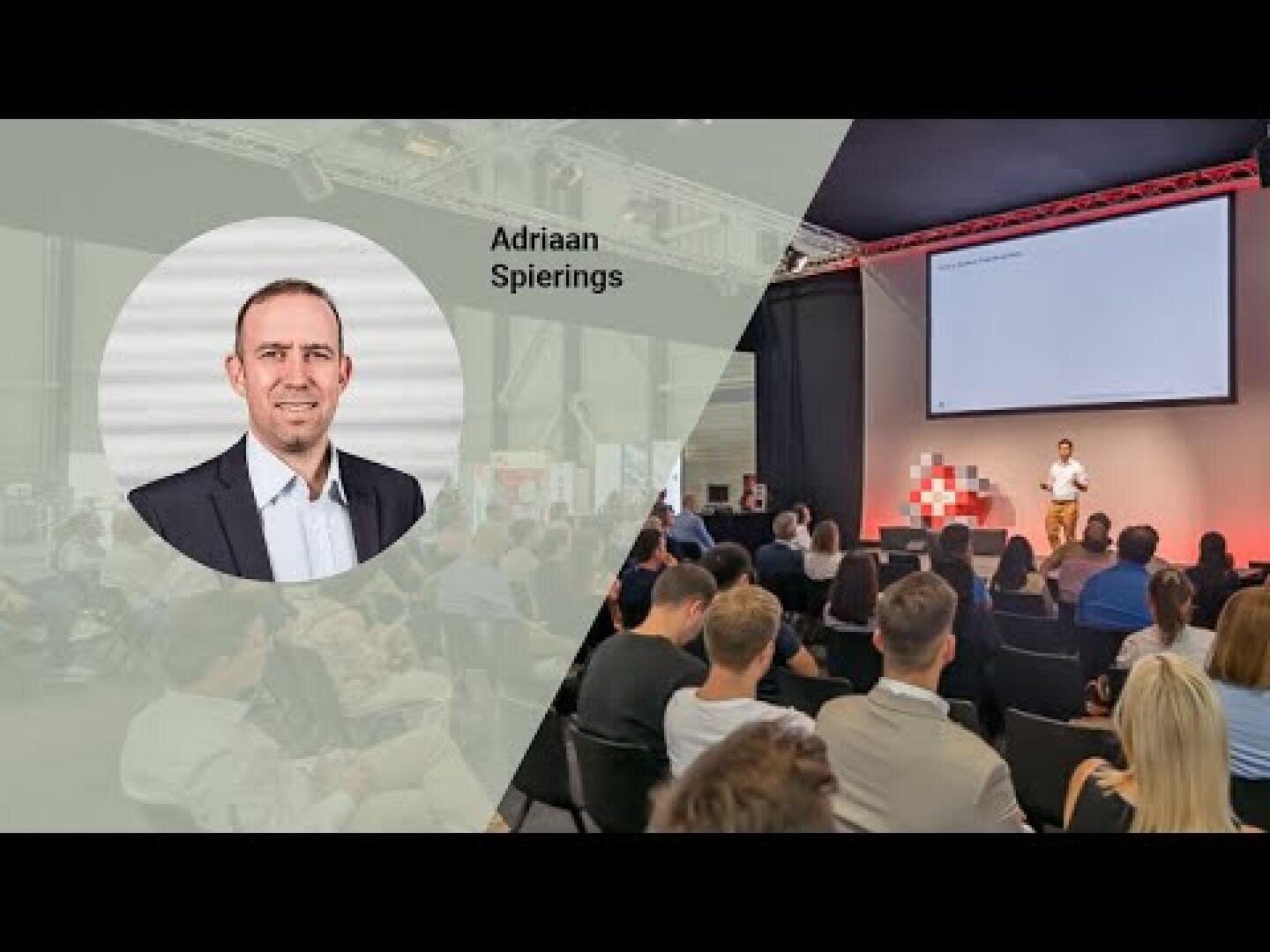 Ein Redner steht auf der Bühne vor einem sitzenden Publikum und präsentiert neben einem großen Bildschirm. Ein transparentes Overlay zeigt ein Kopfbild und den Namen Adriaan Spierings auf der linken Seite des Bildes.