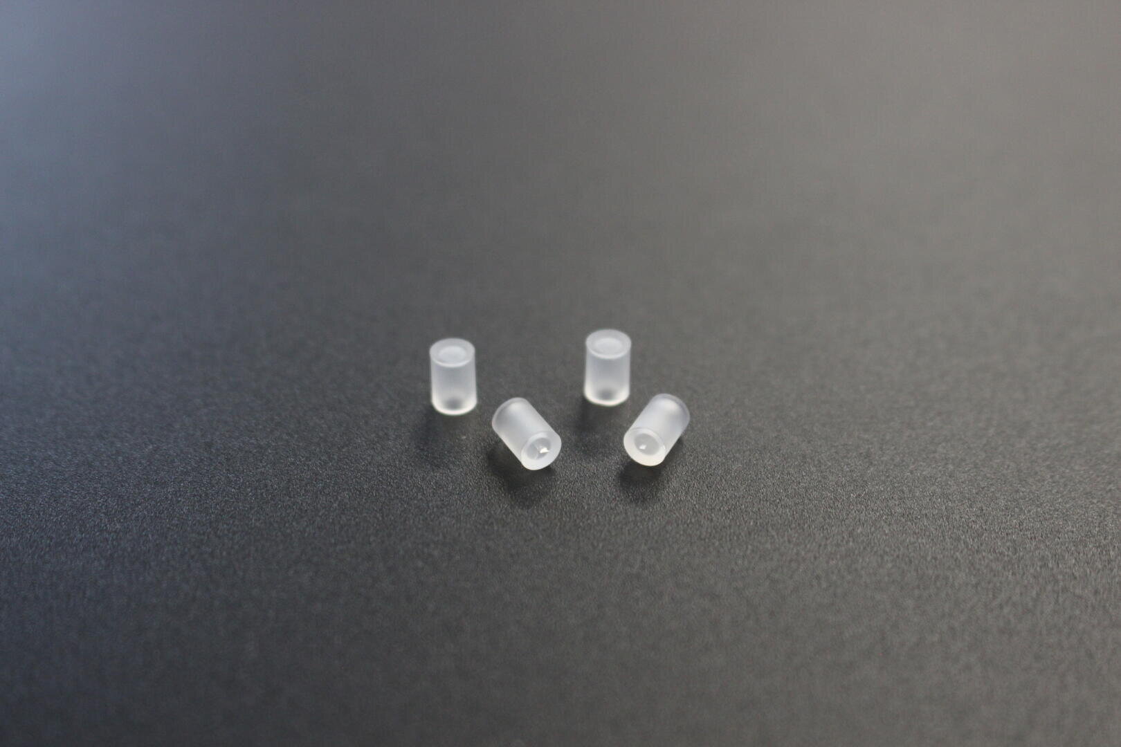 Vier kleine, durchscheinende, zylindrische Kunststoffteile sind auf einer glatten, dunkelgrauen Oberfläche angeordnet.