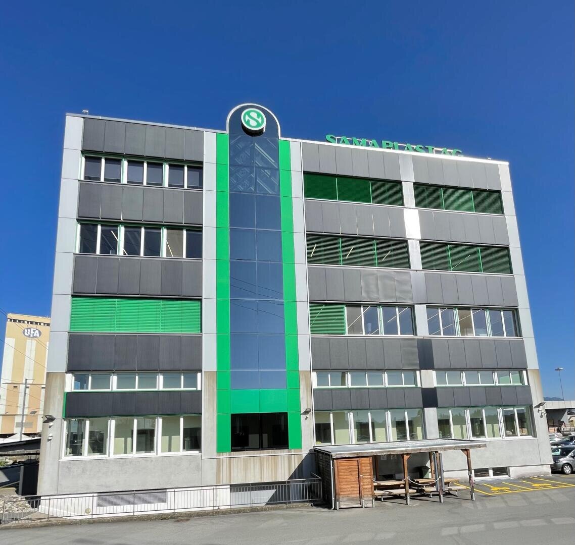 A modern four-story office building with a glass central section and green vertical accents, under a clear blue sky. The sign at the top reads Santa Plast AS. There is a small sheltered area near the entrance.
