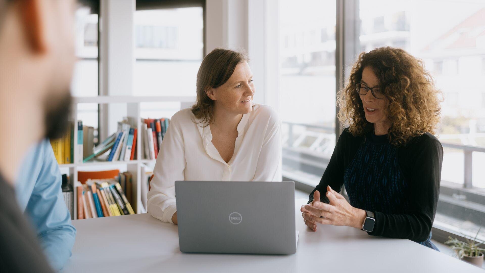 Two women are sitting at a table with a laptop, having a conversation. There are bookshelves and large windows in the background, and two other people are partially visible in the foreground.