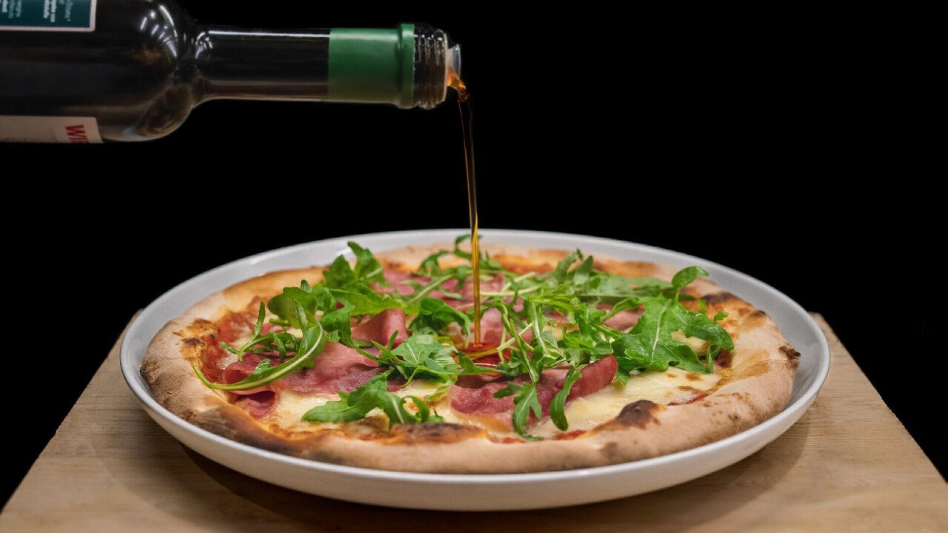 A bottle pours a dark liquid, likely balsamic vinegar or oil, onto a pizza topped with arugula and prosciutto on a white plate against a black background.