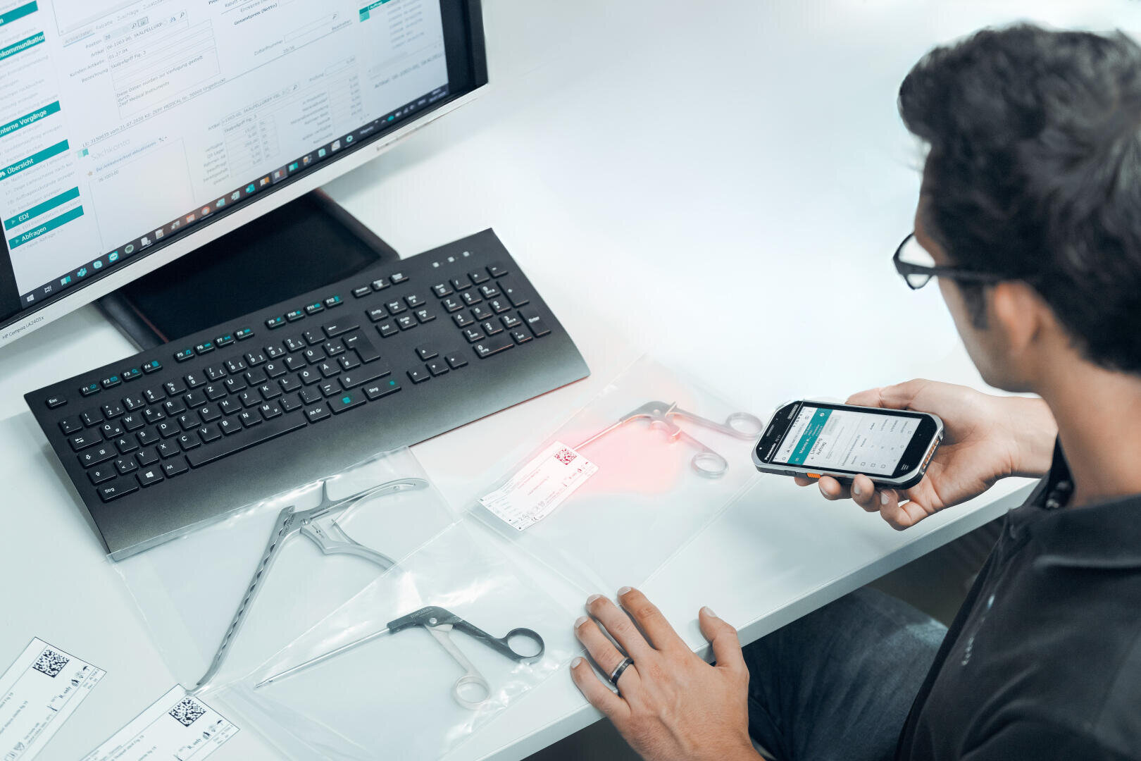 A person sits at a desk with a computer, holding a smartphone and scanning barcodes on medical instruments sealed in plastic bags. Data and forms are displayed on the computer screen.