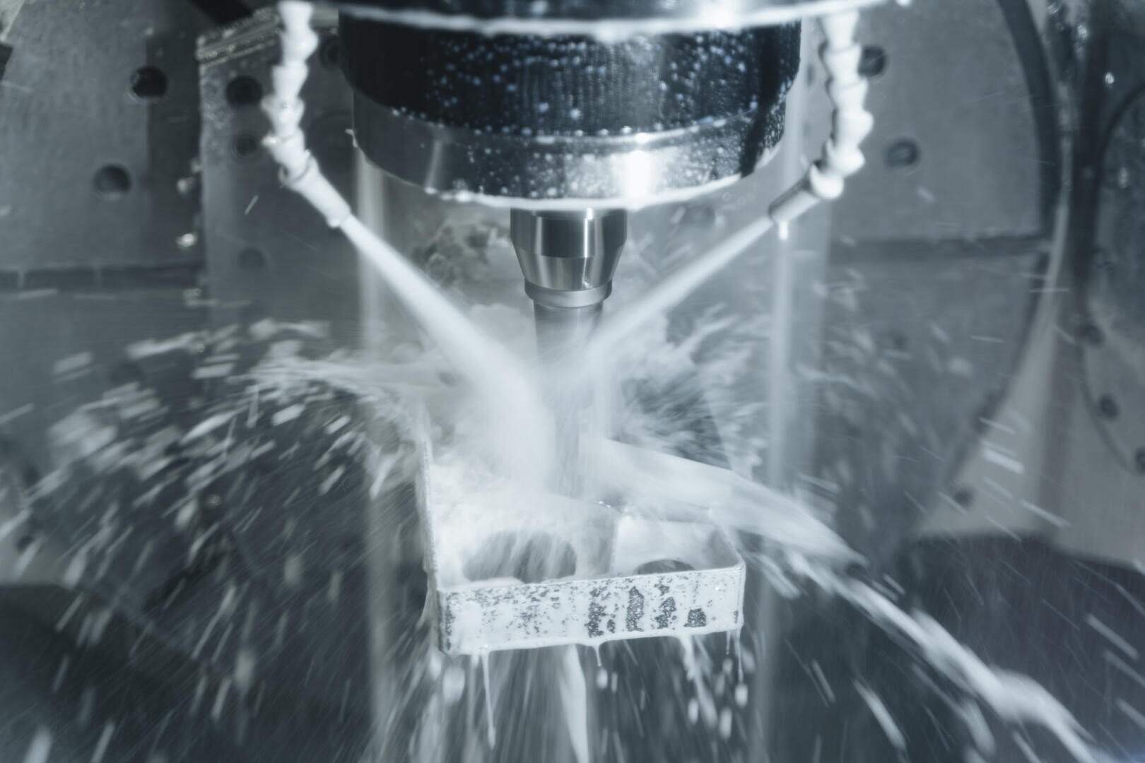A CNC machine processes a metal workpiece with a rotating cutting tool, while coolant sprays surround the tool and create splashes and mist during the milling process.