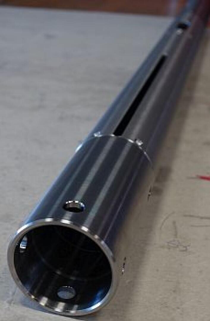 Nahaufnahme eines zylindrischen Metallrohrs mit Löchern und einem langen Schlitz, das auf einer ebenen Fläche liegt. Das Metall erscheint poliert und hat eine reflektierende Oberfläche.