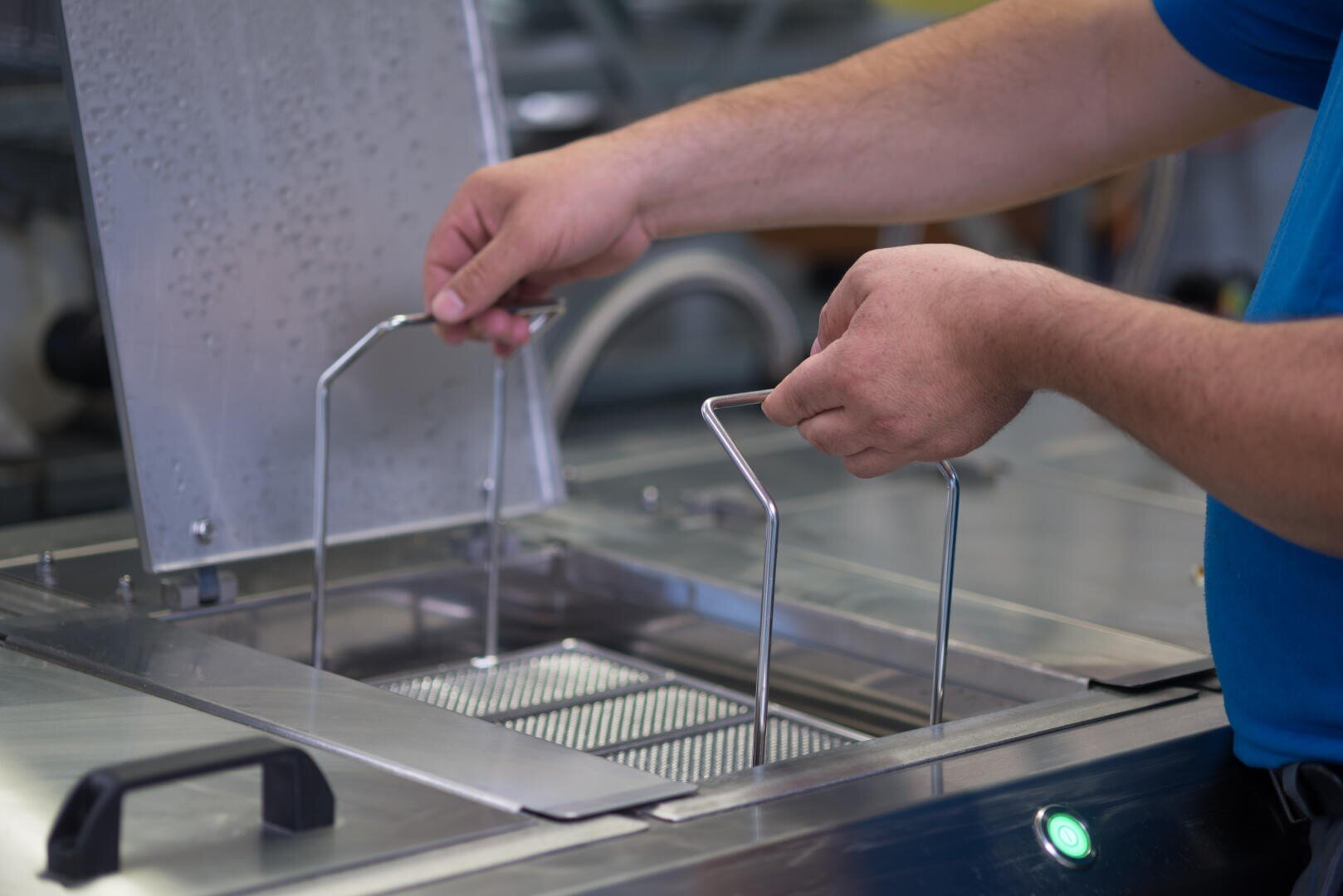 Die Hände einer Person senken einen Metallkorb in eine Industriemaschine aus rostfreiem Stahl, möglicherweise zum Waschen oder Sterilisieren von Geräten, wobei der Deckel der Maschine geöffnet ist.