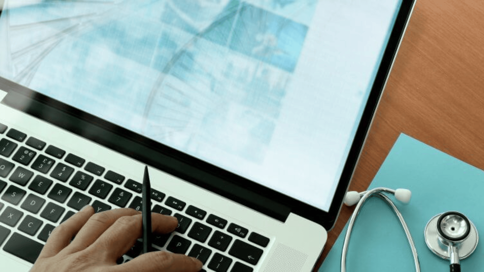 A person's hand uses a pen on a laptop keyboard displaying scientific images, while a stethoscope and a blue folder lie on the wooden table next to the computer.
