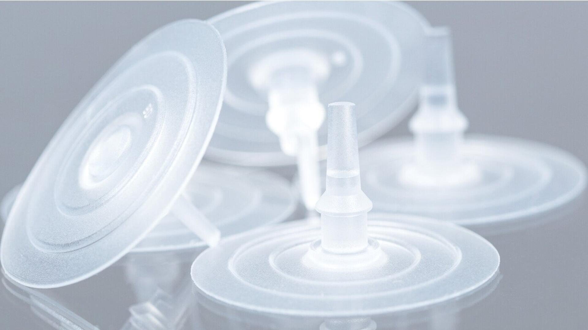 Close-up of several translucent, circular plastic membrane filters with central nozzle-like connectors, arranged overlapping on a reflective gray surface.