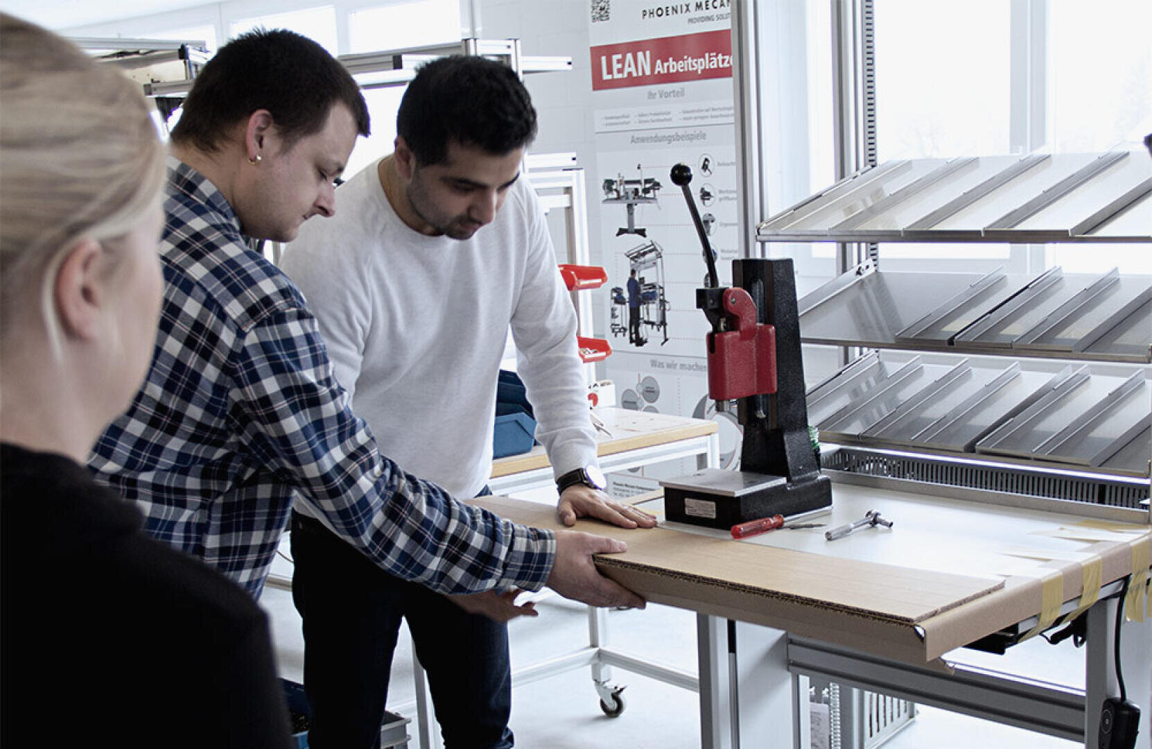 Two men operate a red pressing machine on a workbench while a woman looks on. Behind them are metal shelves, tools and a poster about lean workstations. The surroundings appear to be a workshop or a training room.