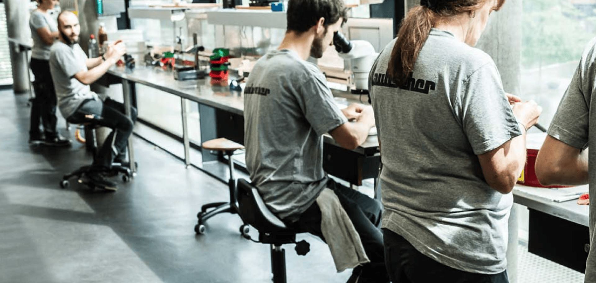 Several people in gray shirts work at long laboratory tables in a bright, modern workroom with large windows. Some are sitting, focused on their tasks, while others are standing and working with equipment.