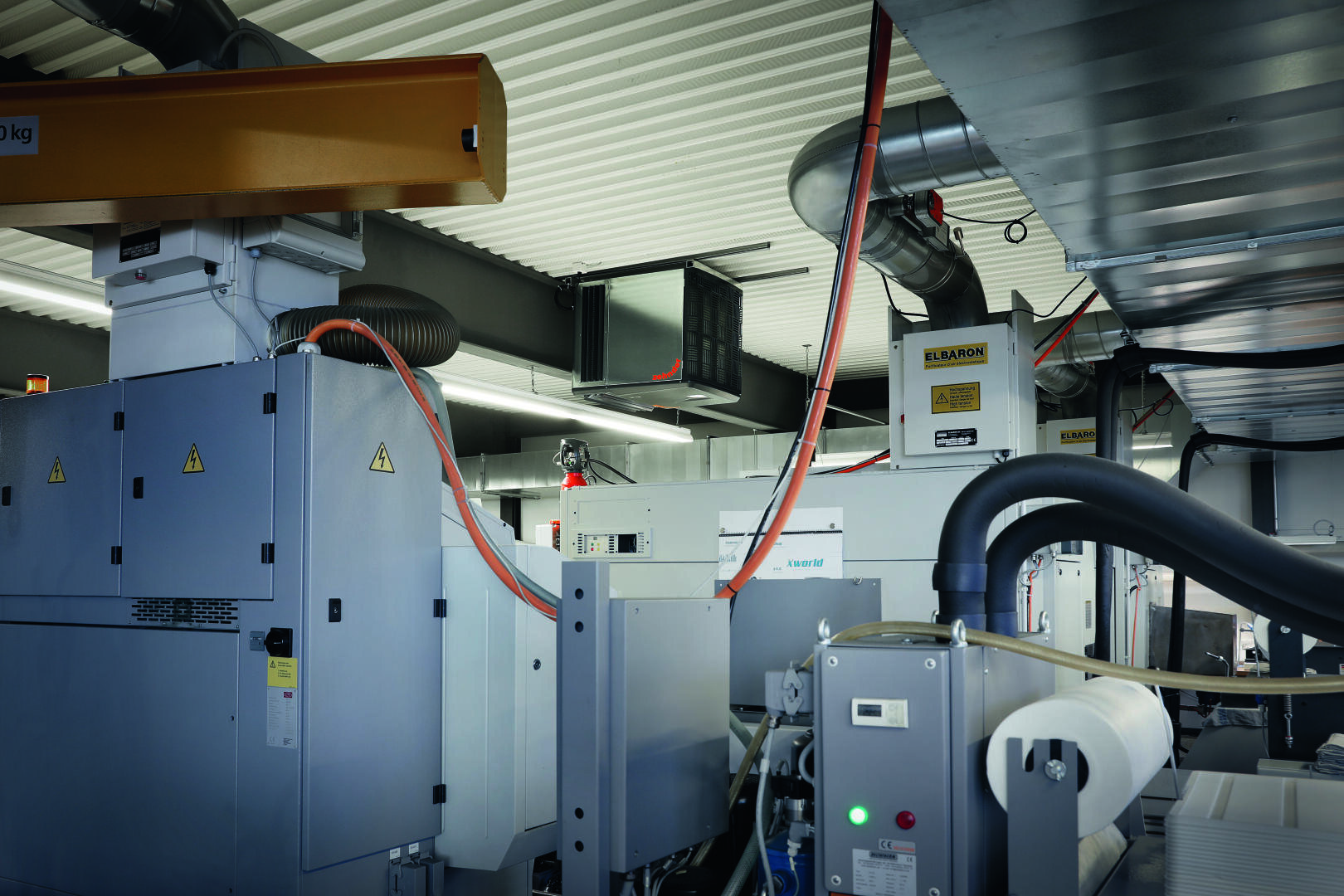 Innenraum einer Industrieanlage mit großen Maschinen, Metallrohren, Schalttafeln und Warnschildern. Über den Köpfen hängen orangefarbene und rote Kabel, und an der Wellblechdecke sind Leuchtstoffröhren angebracht.