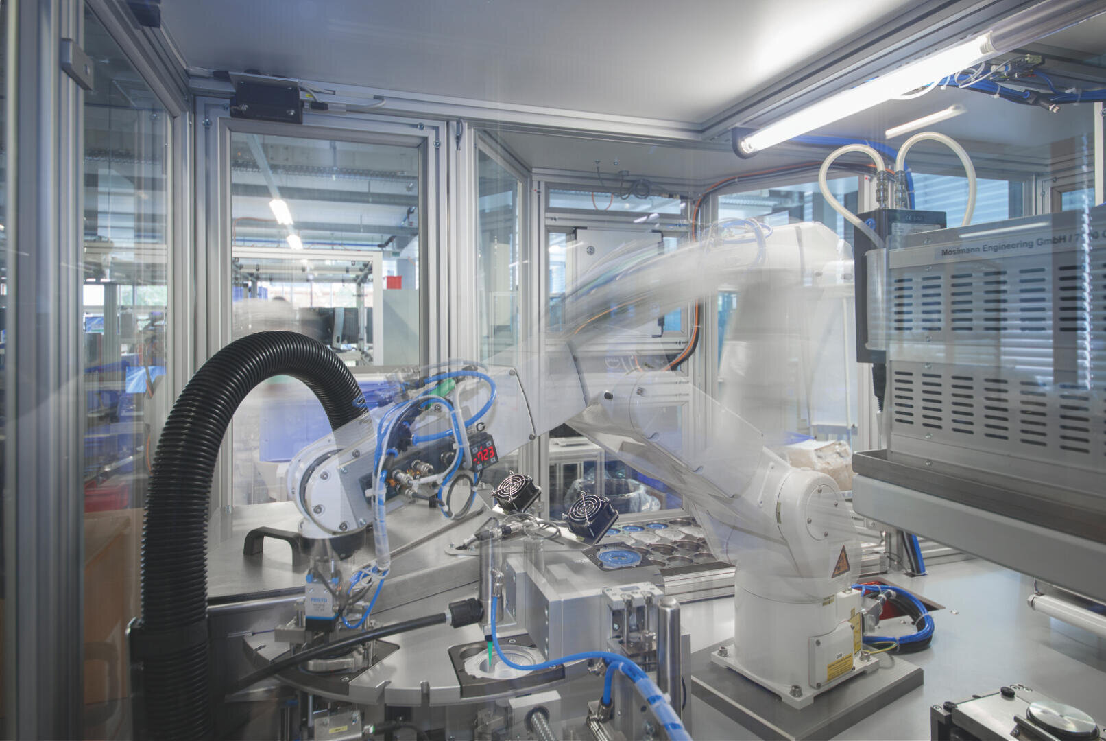 A robotic arm works in a modern industrial laboratory with automated machines and electronic components behind glass panes, while various cables and devices can be seen in the background.