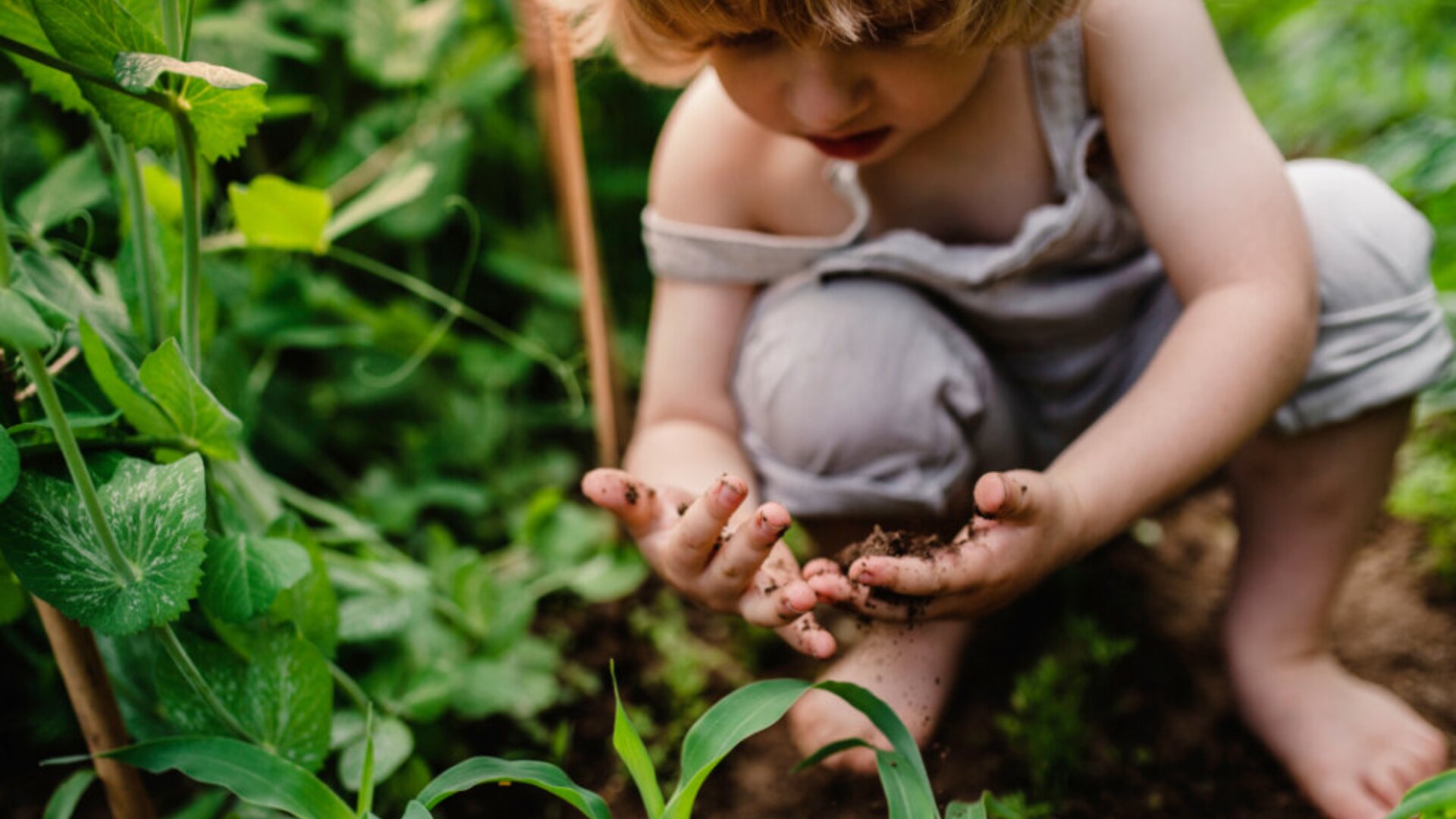 Ein kleines Kind hockt barfuß in einem Garten und untersucht die Erde mit seinen schmutzigen Händen. Grüne Pflanzen umgeben es, während es konzentriert auf den Boden schaut.