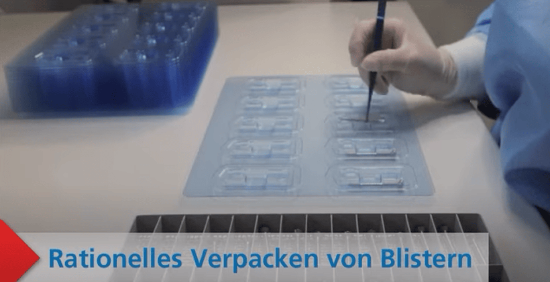 Eine Person in Schutzkleidung arbeitet mit einem Werkzeug an einer durchsichtigen Kunststoffschale für Blisterverpackungen auf einem Tisch. Der deutsche Text am unteren Rand lautet Rationelles Verpacken von Blistern.