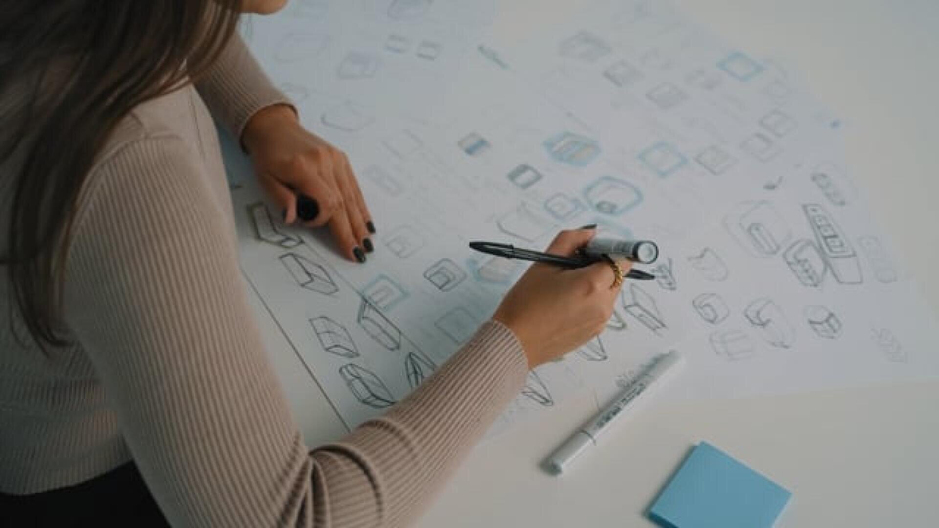 A person sketching various geometric 3D shapes on large sheets of paper with a black marker; on the desk are further drawings, a white marker and blue sticky notes.