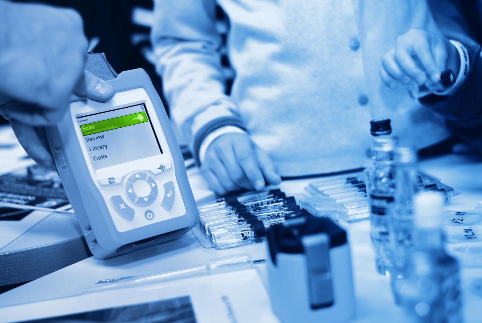 Close-up of hands operating a hand-held electronic device with a screen displaying menu options, surrounded by laboratory equipment and small vials on a table. The scene is colored blue.