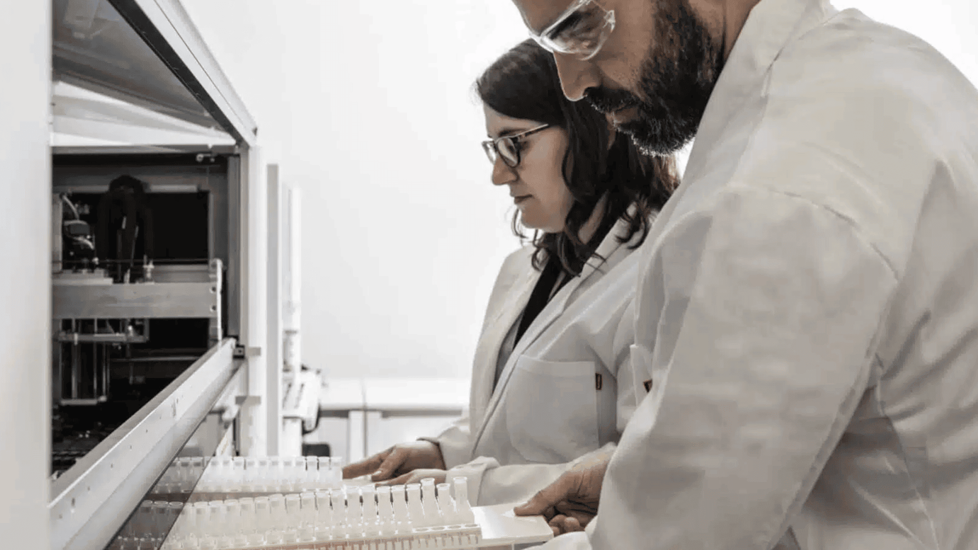 Two scientists in lab coats and protective goggles work with test tubes and laboratory equipment in a modern laboratory environment.