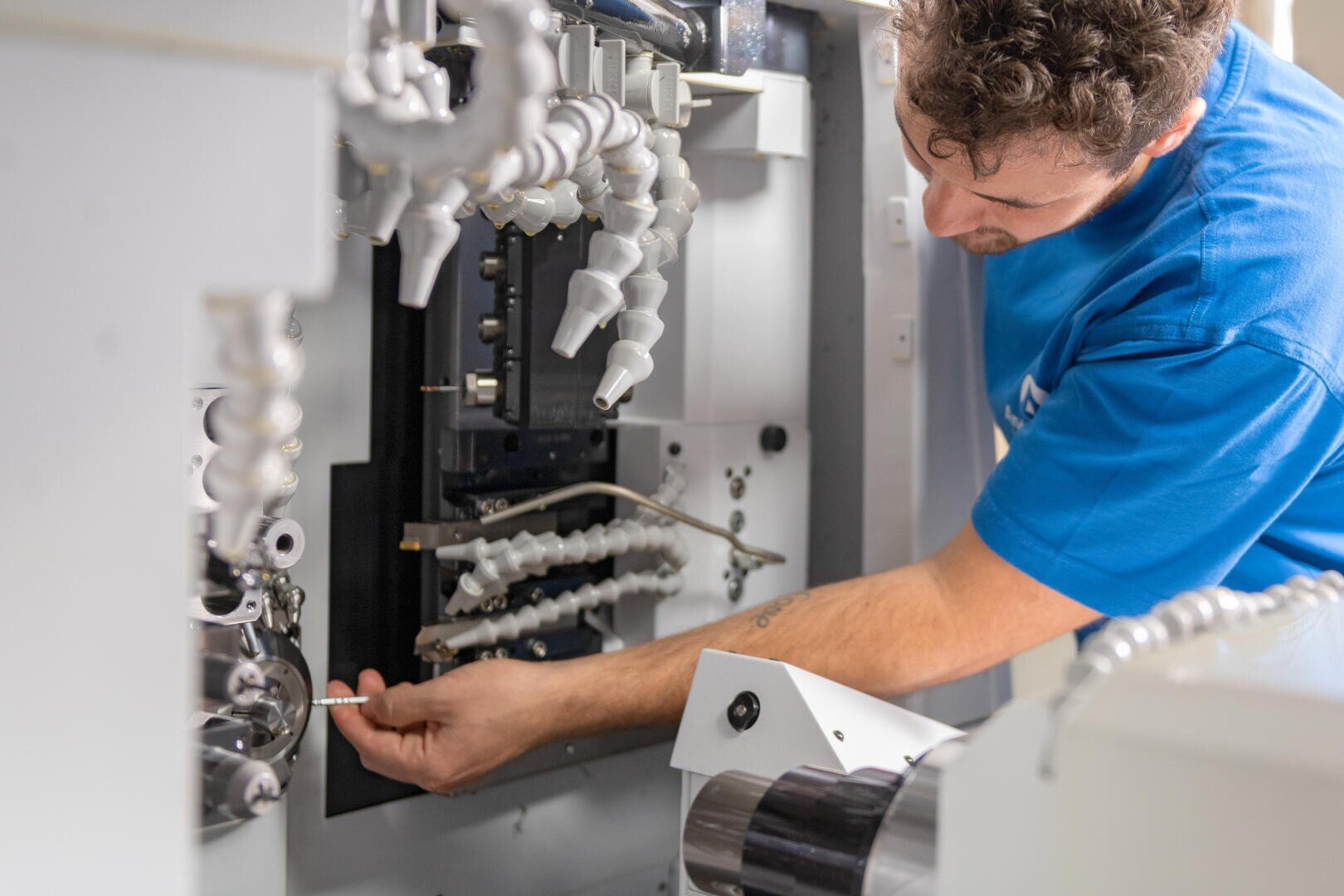 Ein Mann in einem blauen Hemd justiert oder repariert Komponenten in einer Industriemaschine, indem er mit beiden Händen ein Werkzeug benutzt. In der Maschine sind mehrere weiße flexible Schläuche und Metallteile zu sehen.