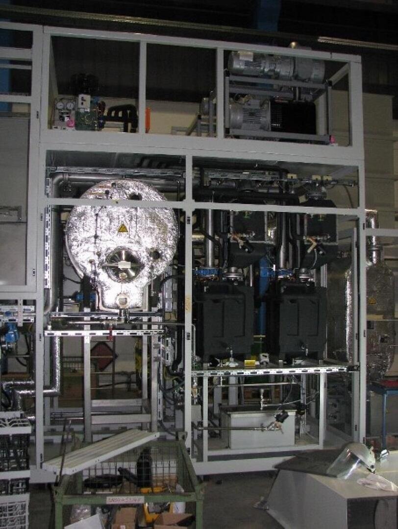 Große Industriemaschine mit freiliegenden Bauteilen, einschließlich Rohren, Tanks und einer runden, mit Folie überzogenen Kammer, die in einem Metallrahmen in einer Fabrik oder einem Labor untergebracht ist.