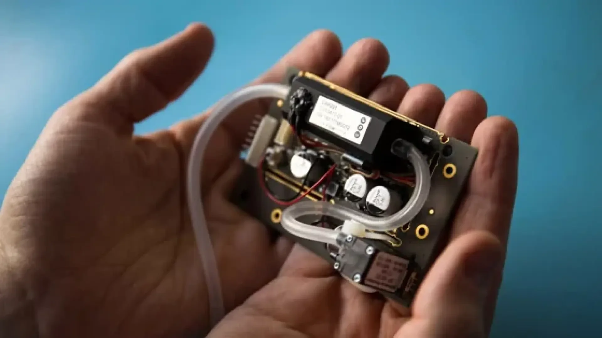 A person holds a small electronic device with wires, circuits and transparent tubes in front of a blue background, suggesting that it is a piece of advanced technology or a prototype.