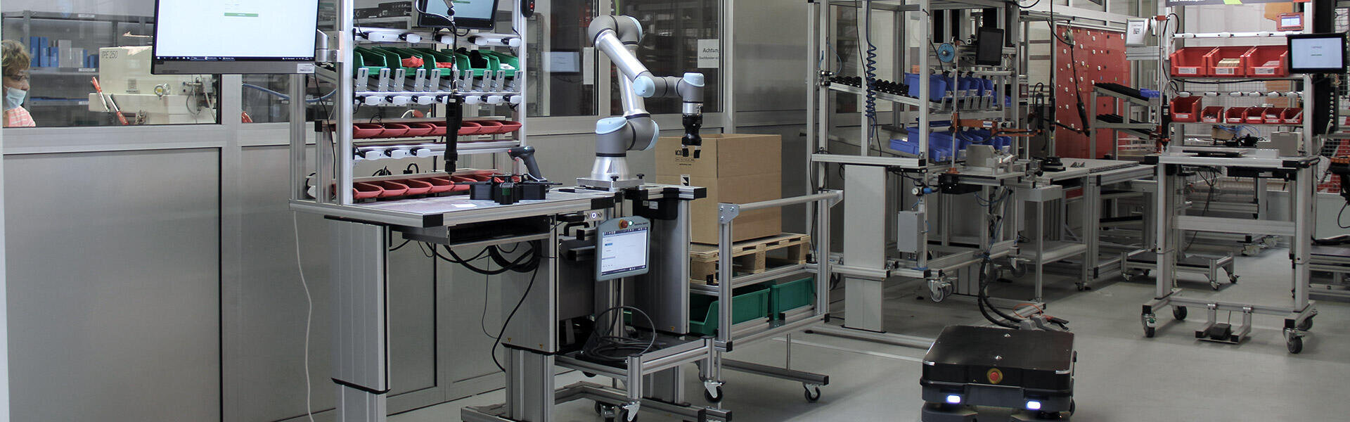 Ein Roboterarm arbeitet an einem Arbeitsplatz mit organisierten Teilebehältern, einem Monitor und einem mobilen Roboter auf dem Boden in einer industriellen, automatisierten Fabrikumgebung.