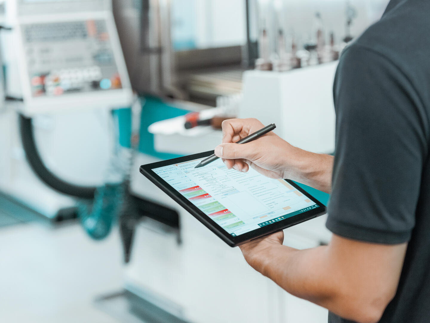 A person is holding a tablet and entering data with a stylus. Blurred industrial machinery can be seen in the background, reminiscent of a high-tech production environment or a technical setting.