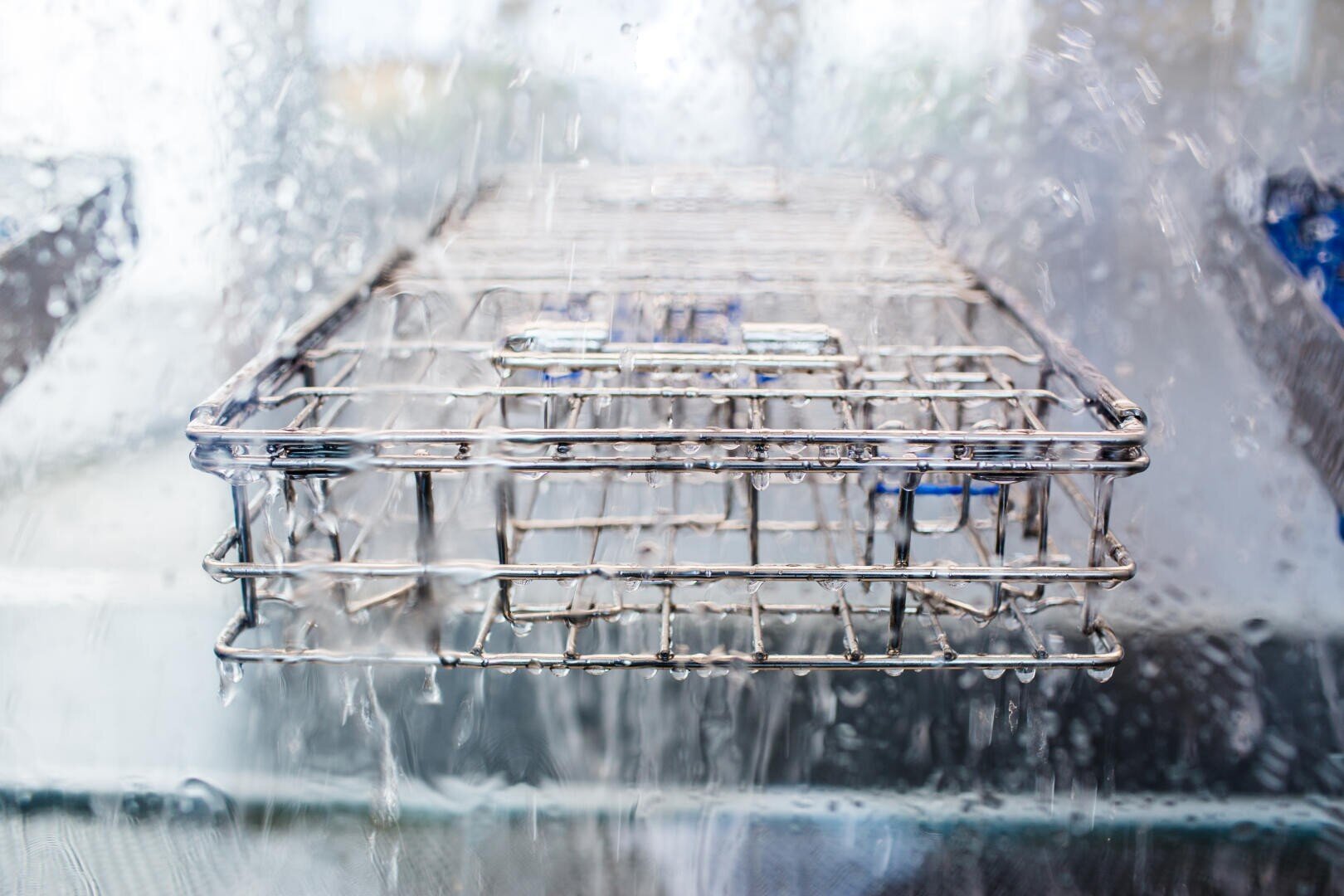 Close-up of a metal rack being sprayed with water inside a washing or sterilizing machine, with water droplets and splashes visible on the glass surface.