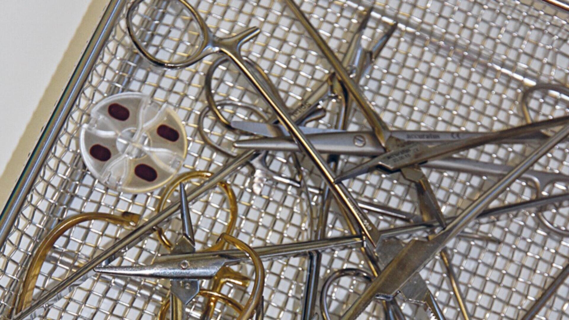 A metal tray containing various surgical instruments, including scissors, forceps, and hemostats, with a small round object with four dark areas also visible.
