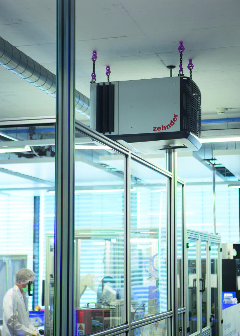 A lab worker in protective gear works inside a glass-enclosed laboratory, while a Zehnder-branded air purifier is suspended from the ceiling above.