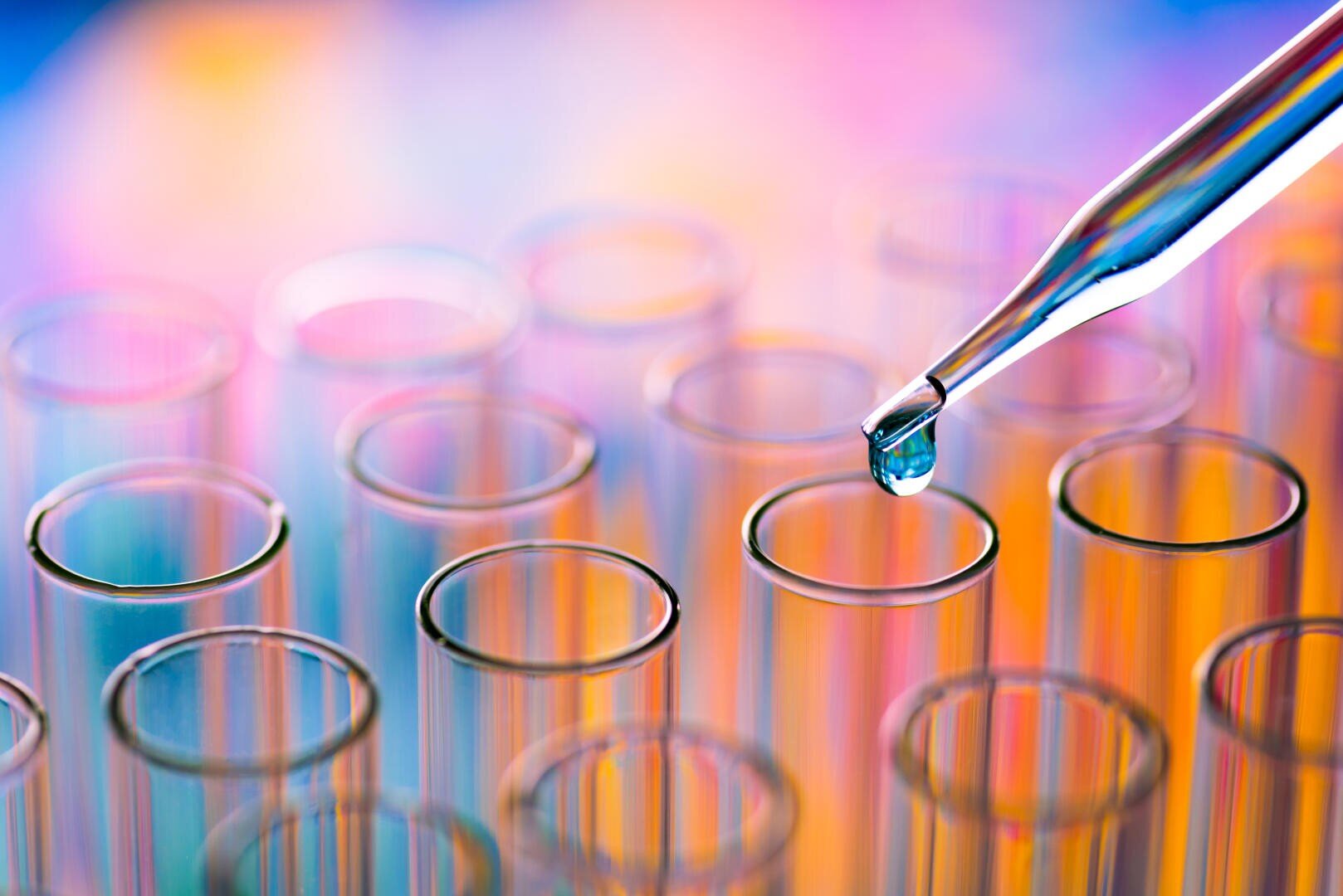 Close-up of a pipette dispensing a drop of liquid into one of several transparent test tubes, with a colorful, blurred background.