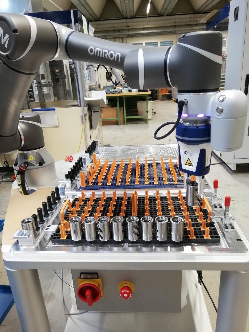 A robotic arm labeled Omron works over a workbench with organized metal parts and orange plastic holders in an industrial setting, suggesting automated assembly or sorting.