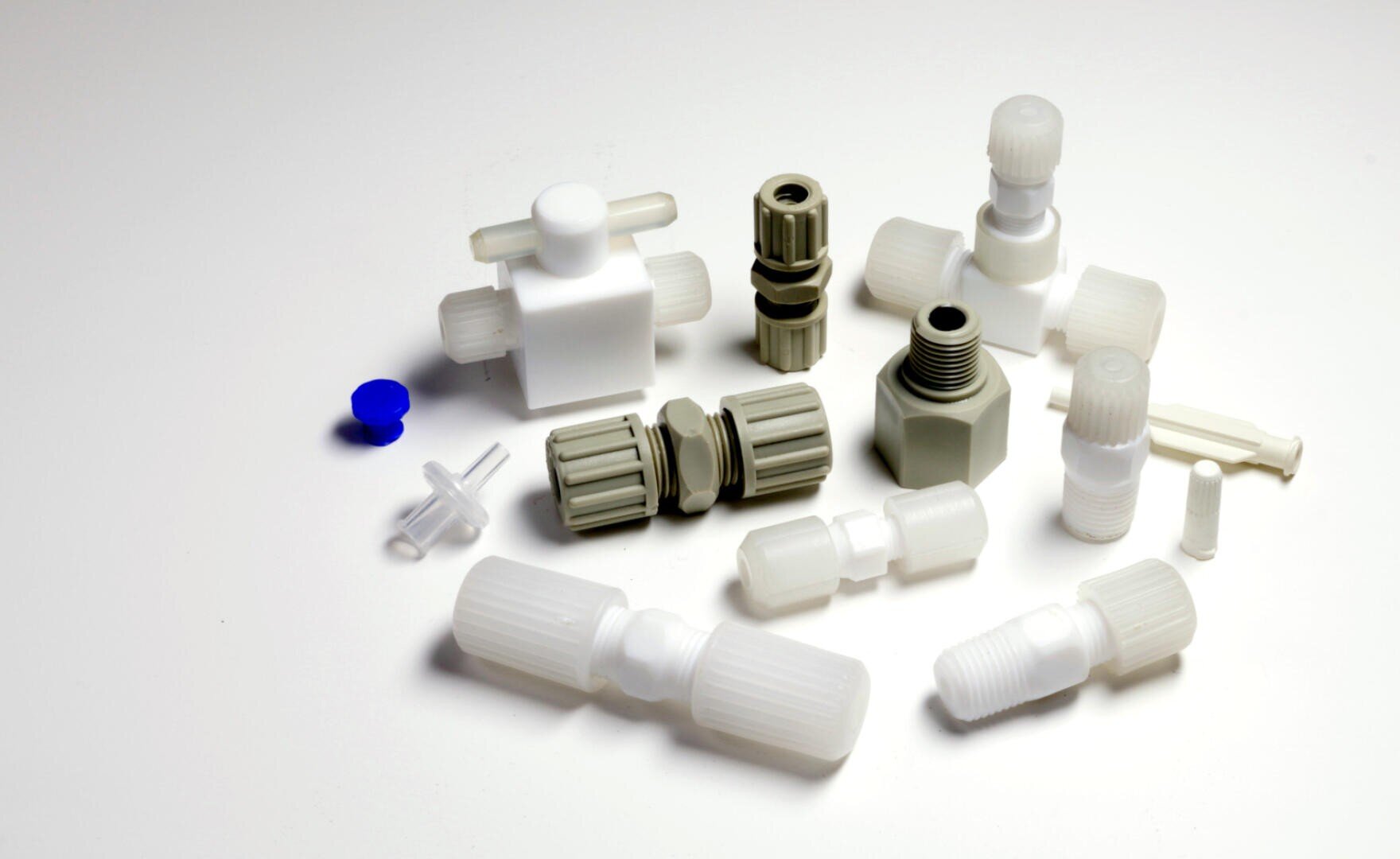 Auf einer weißen Fläche sind verschiedene weiße und graue Kunststoffbeschläge, Verbindungsstücke und Kappen angeordnet, darunter auch Teile mit Gewinde und Rillen, darunter eine kleine blaue Kappe.