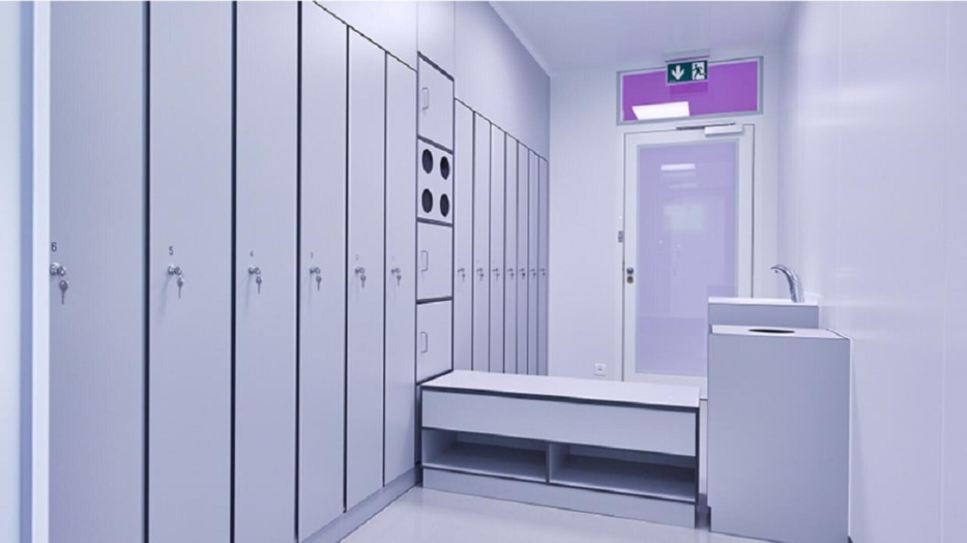 Ein moderner Umkleideraum mit hellgrauen Spinden, einer Bank, einem Waschbecken und einer Ausgangstür mit einem grünen Notfallschild darüber. Der Raum ist sauber, minimalistisch und durch Oberlichter hell erleuchtet.