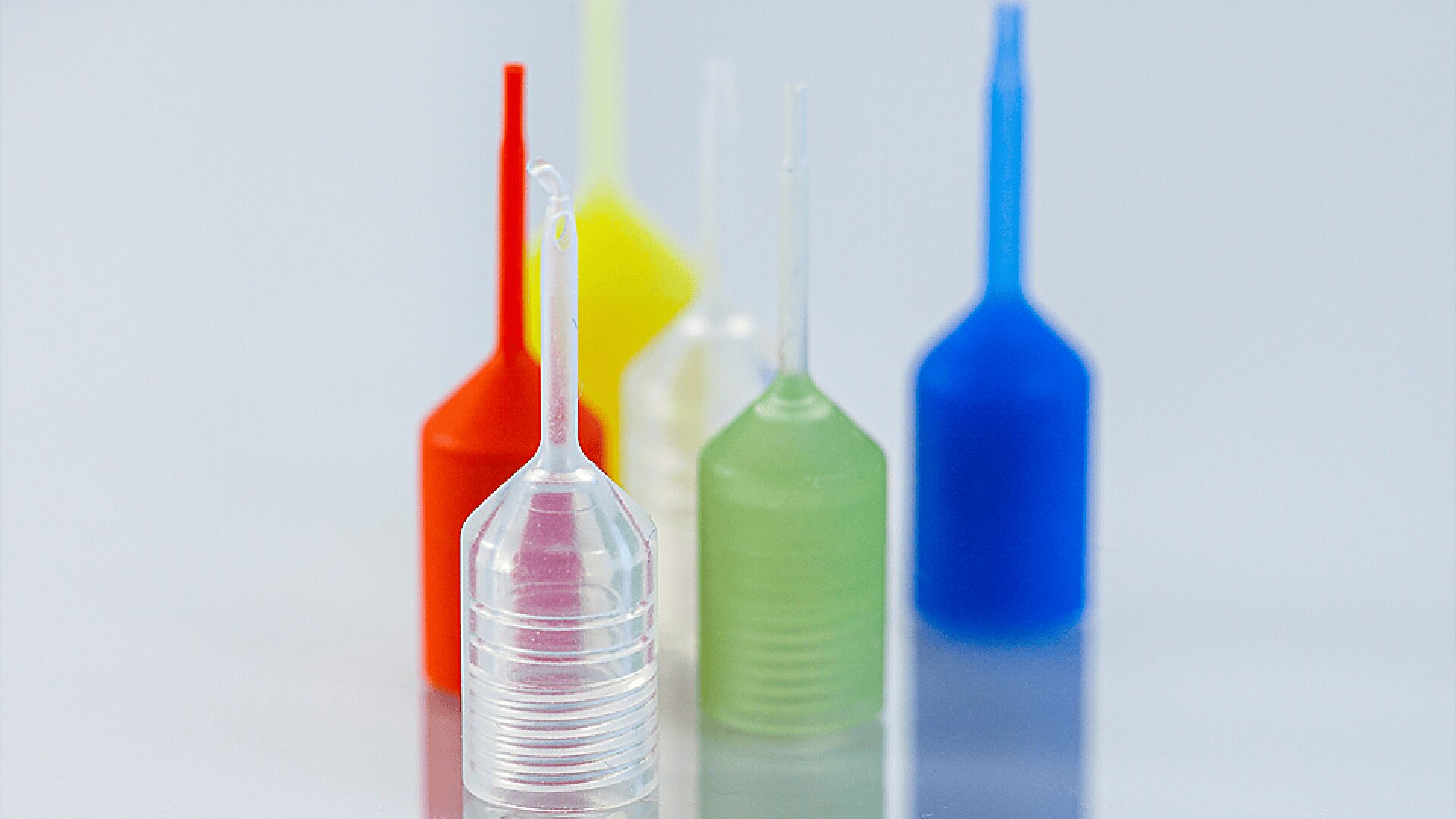 Fünf kleine, bunte Plastikdüsen stehen aufrecht auf einer weißen Fläche. Die Düsen sind rot, gelb, klar, grün und blau und mit Ausnahme der klaren Düse in der Mitte leicht unscharf.