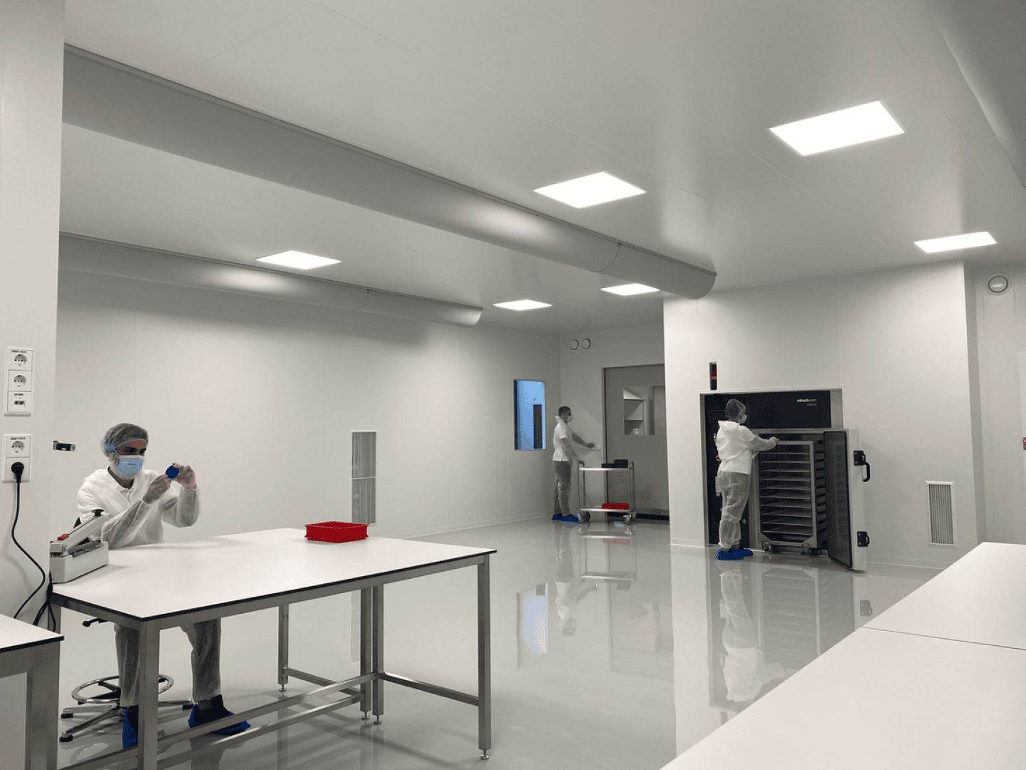 Three people in full protective suits work in a clean, white laboratory. One sits at a table handling equipment, while two others stand near large machinery. The room is bright and sterile with minimal furnishings.