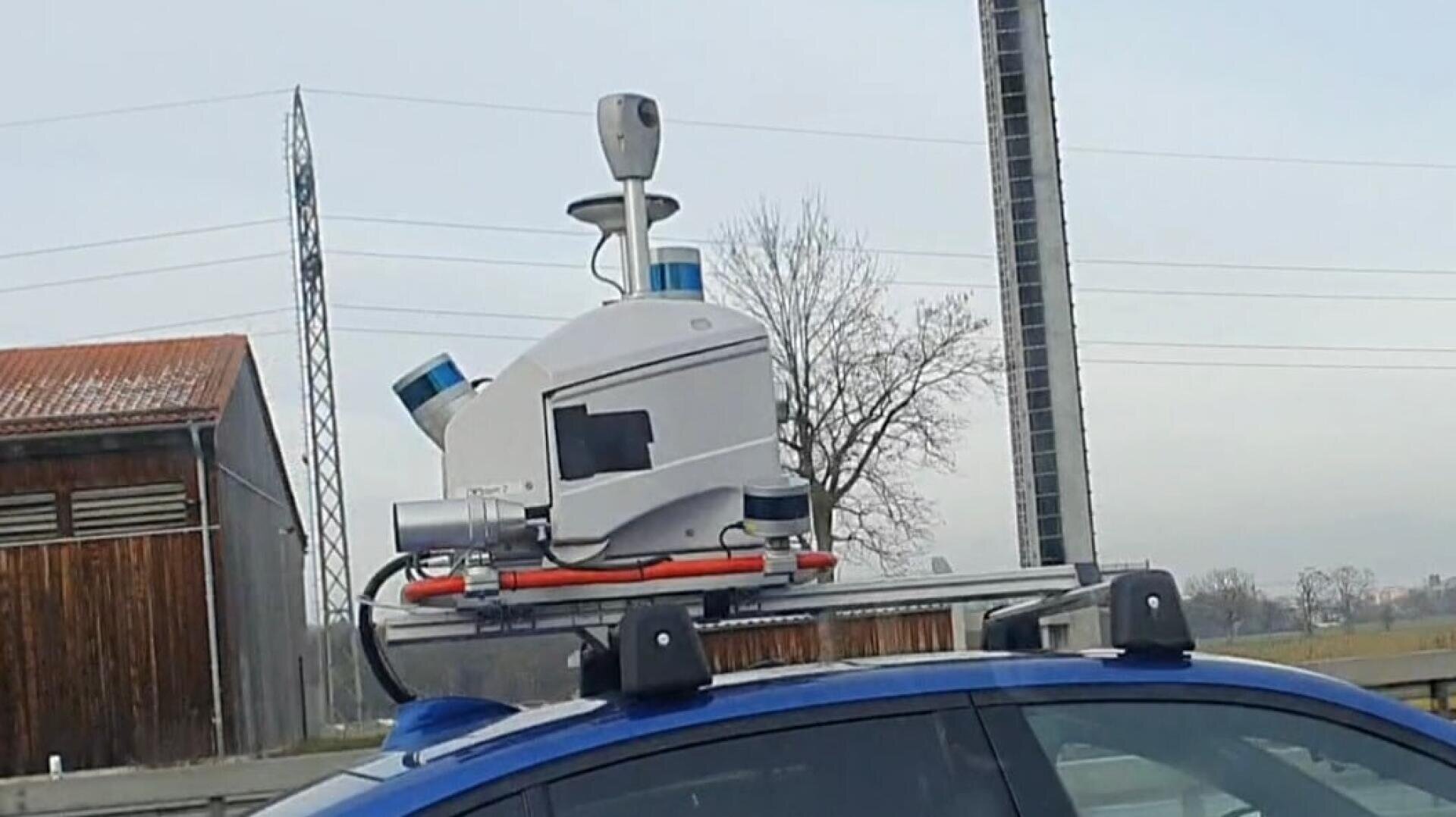 Ein blaues Auto, auf dessen Dach ein komplexes Sensorsystem mit Kameras, Lidarsensoren und anderen Geräten montiert ist, das wahrscheinlich für die Kartierung oder die Erforschung des autonomen Fahrens verwendet wird. Im Hintergrund sind ein Holzgebäude, ein Mast und Bäume zu sehen.