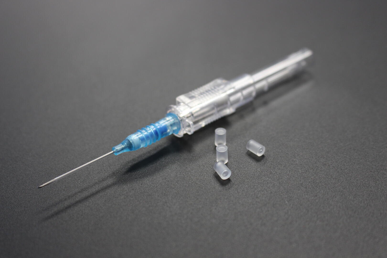 Nahaufnahme einer medizinischen Spritze mit Nadel, mit transparentem Zylinder und blauen Akzenten, die auf einer dunklen Fläche neben drei kleinen, klaren Plastikkappen liegt.